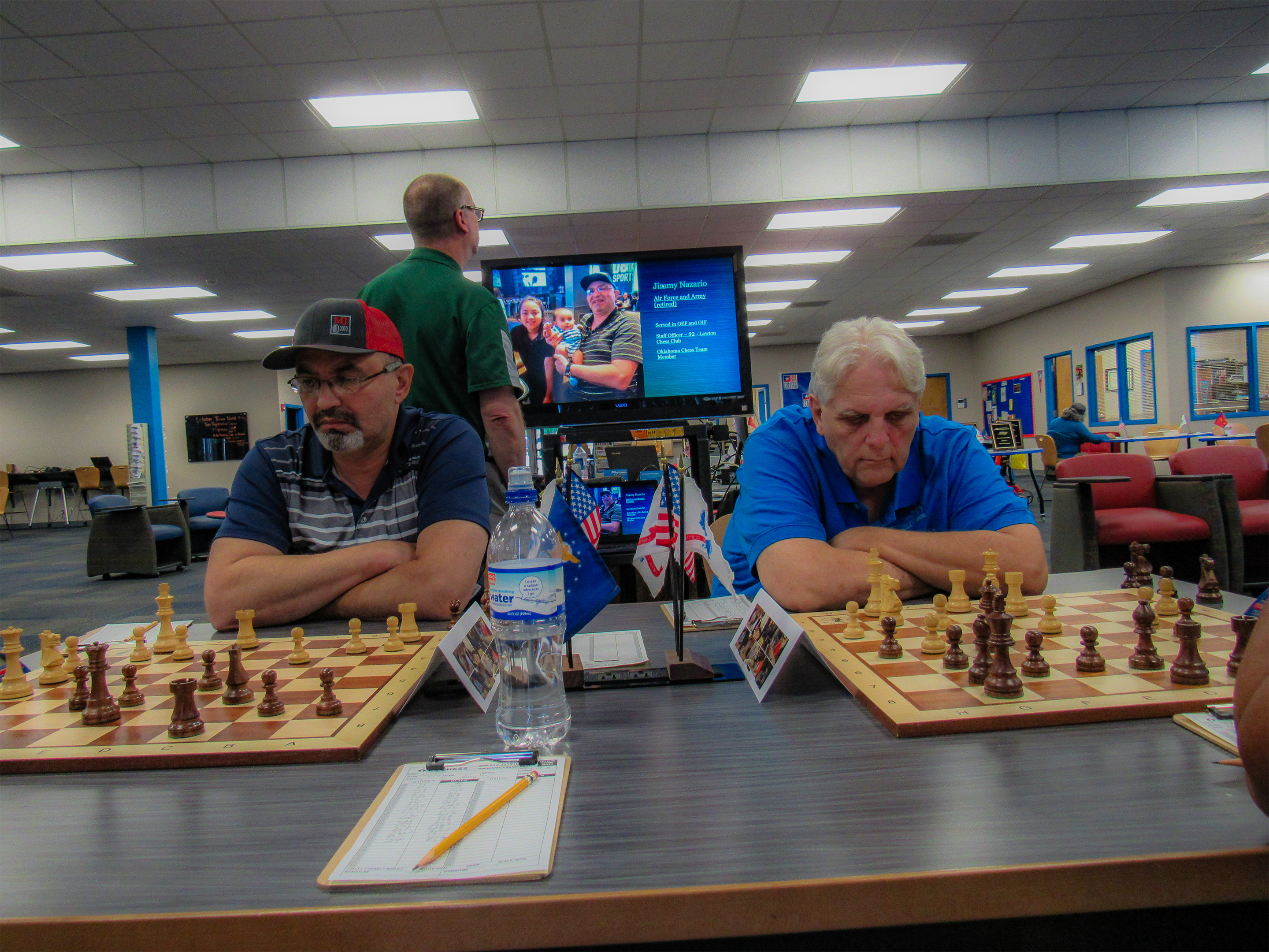 Air Force/Army Retiree Jimmy Nazario (left) and Army Veteran Troy Gillispie (right) doing battle side-by-side.  In the background Chess Expert Ron Farrar observes a continuously running slideshow about this Championship.  Photo by Sheryl McBroom.